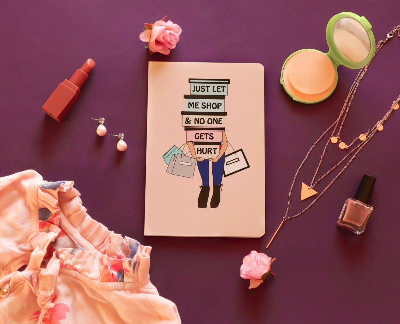 A notebook with the phrase 'JUST LET ME SHOP & NO ONE GETS HURT' printed on the cover, presented on a purple background with fashion-related items like clothes, earrings, and a handbag.