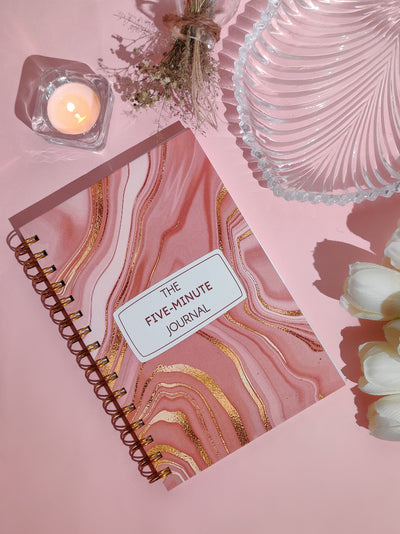 Pink marbled journal with 'The Five-Minute Journal' on a pink background with decorative items.